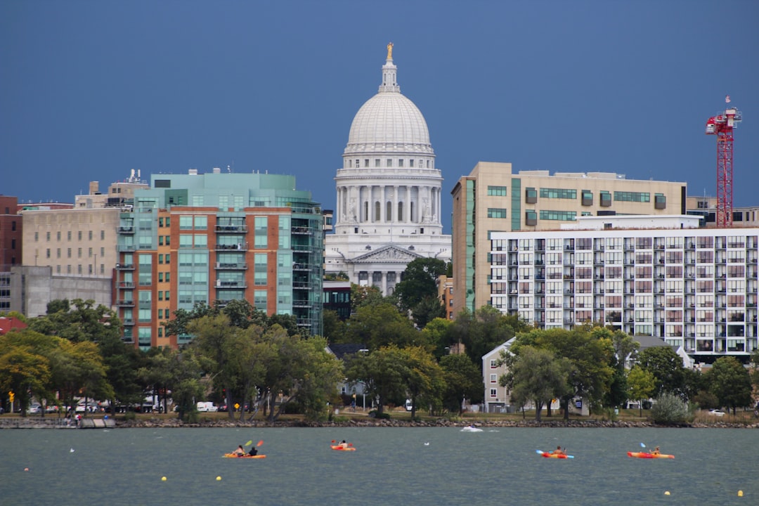 The Wisconsin State Capitol building dominates the skyline of Madison, Wisconsin. The iconic dome is visible above surrounding skyscrapers and trees. People are kayaking in Lake Monona in the foreground.