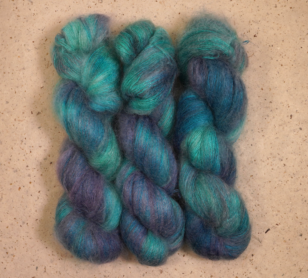 Tres madejas torcidas de hilo de mohair jaspeado en verde azulado, azul y morado, sobre una superficie de piedra beige texturizada