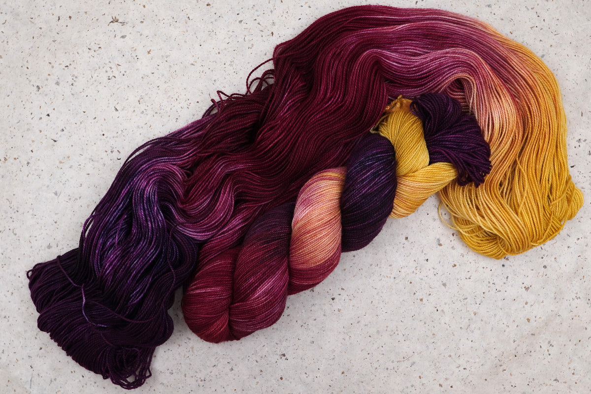 Hilo Maizie Dayz de Wild Woollys — una madeja abierta en morado intenso, dorado, magenta y carmesí