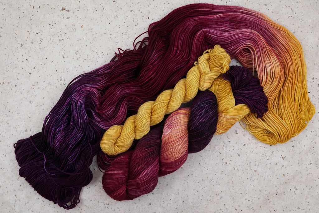 Wild Woollys Maizie Dayz sock set, an unwound skein in deep purple, gold, magenta, and crimson with a contrasting golden yellow mini-skein