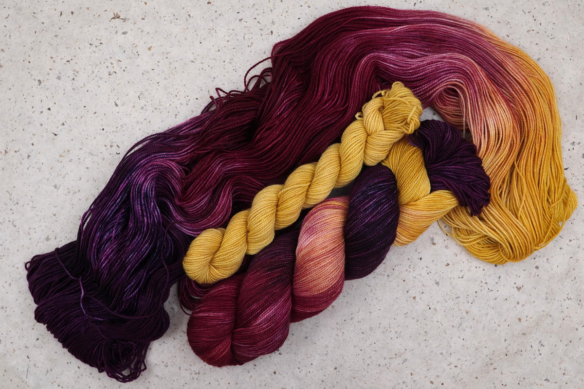 Wild Woollys Maizie Dayz sock set, an unwound skein in deep purple, gold, magenta, and crimson with a contrasting golden yellow mini-skein
