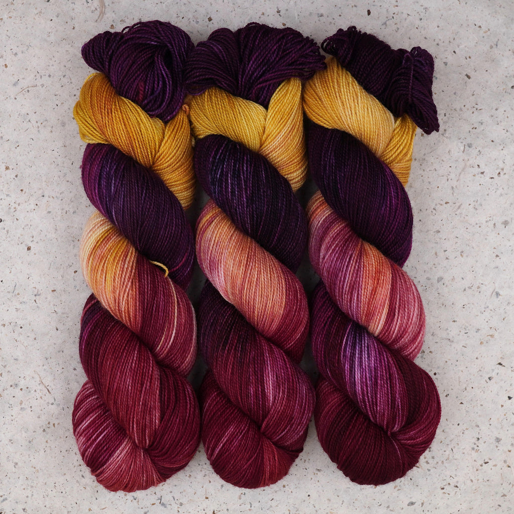 Wild Woollys Maizie Dayz Fingering yarn, three skeins in deep purple, gold, magenta, and crimson