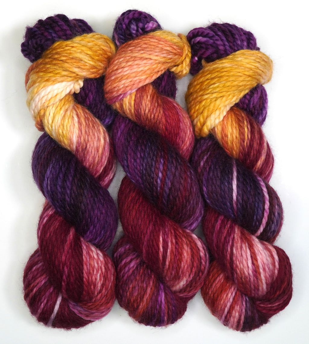 Three skeins of Wild Woollys Maizie Dayz yarn in shades of purple, orange, and red