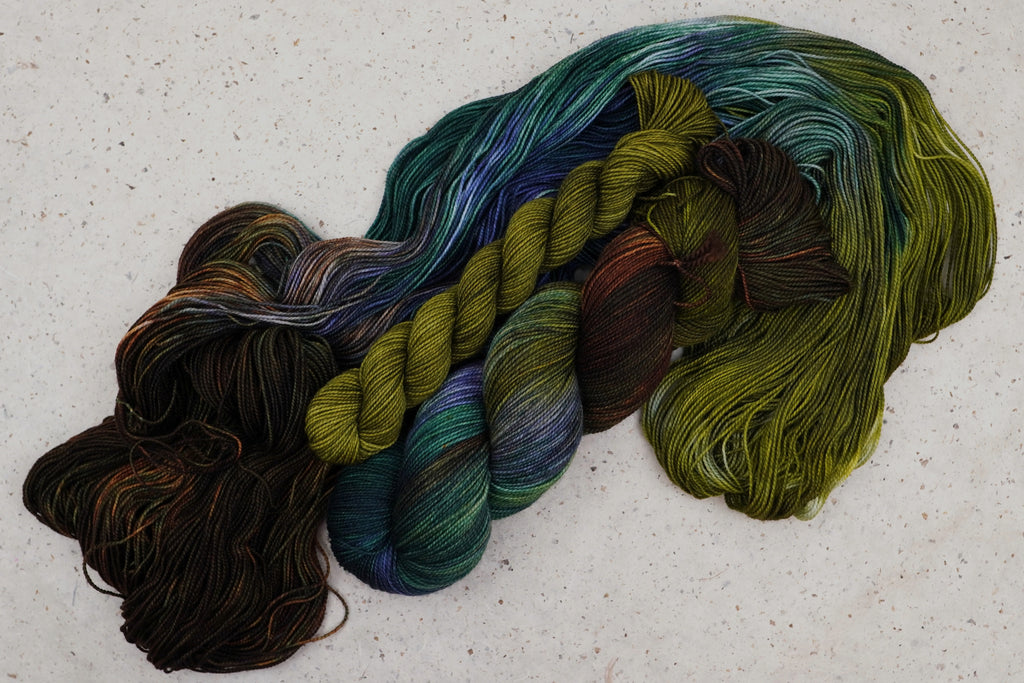 Kit chaussettes Mycelium Bloom de Wild Woollys — un écheveau déroulé aux tons vert olive, brun foncé et bleu canard profond avec une mini-pelote contrastante doré olive