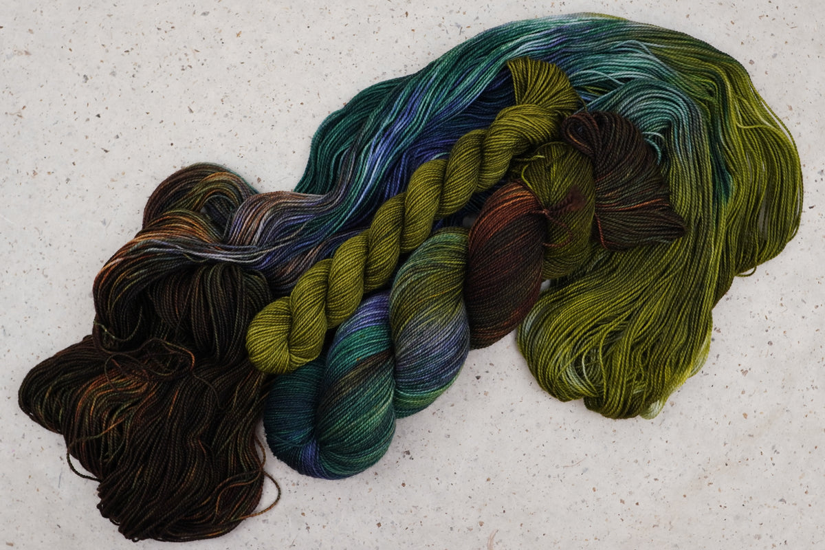 Wild Woollys Mycelium Bloom sock set, an unwound skein in olive green, dark brown, and deep teal with a contrasting olive gold mini-skein