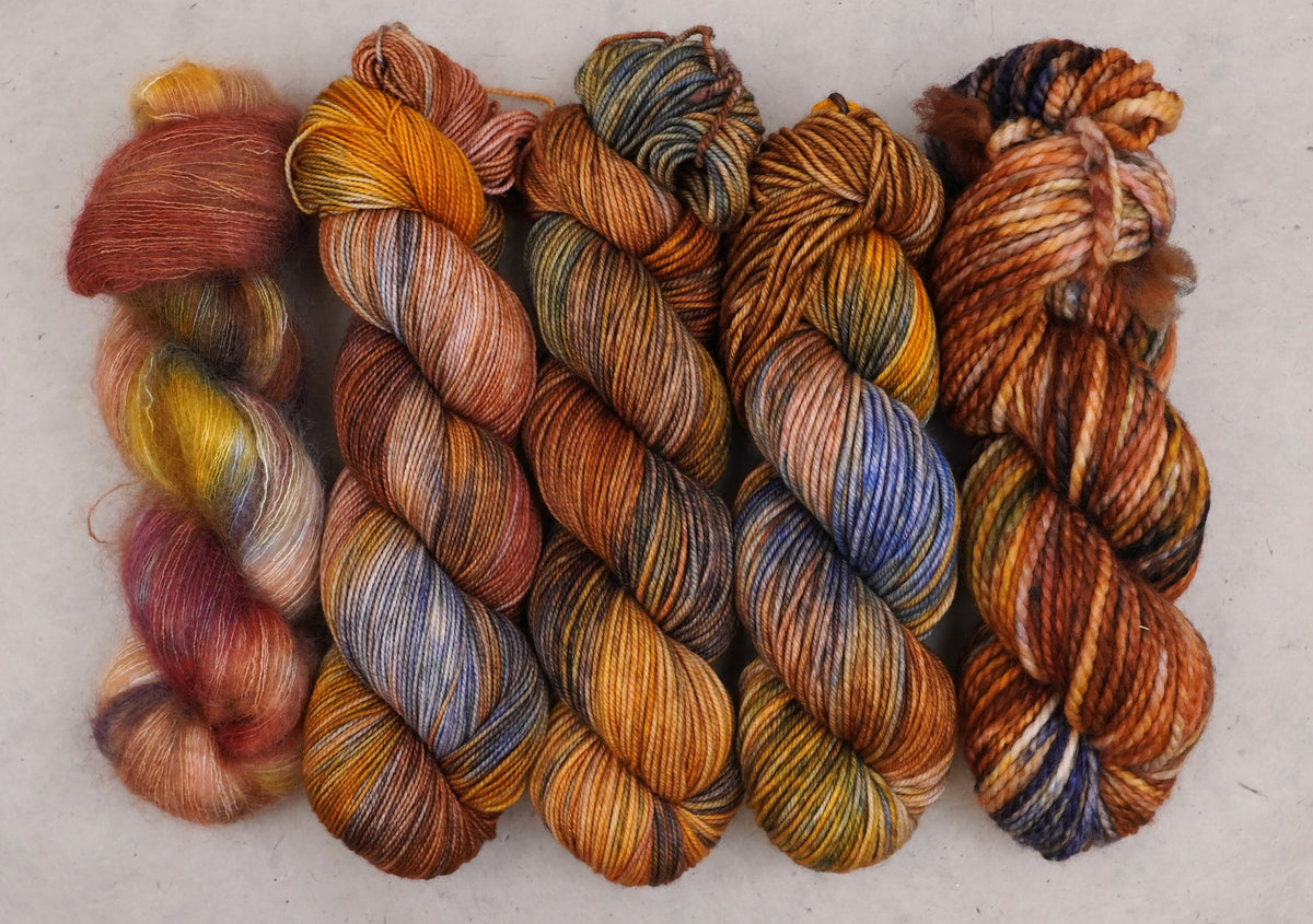 Six skeins of variegated yarn arranged side by side on a light neutral background, showcasing a mix of warm and cool colors including orange, brown, yellow, blue, and red.