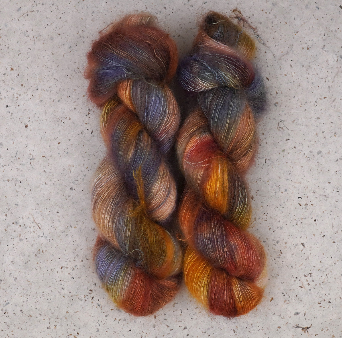 Two twisted hanks of variegated yarn laid flat on a light speckled stone surface. The yarn displays a blend of warm rust orange, golden yellow, peach, and muted blue-gray tones.