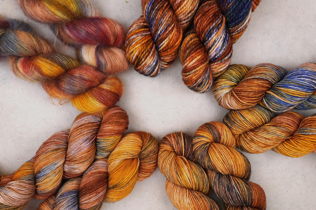Multiple twisted skeins of variegated yarn arranged on a light neutral background, showing blended warm and cool colors including orange, brown, gold, blue, purple, and cream.
