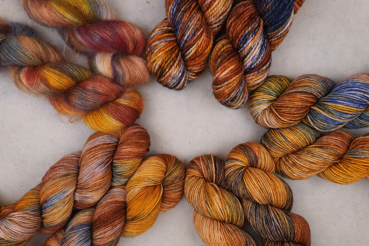 Multiple twisted skeins of variegated yarn arranged on a light neutral background, showing blended warm and cool colors including orange, brown, gold, blue, purple, and cream.