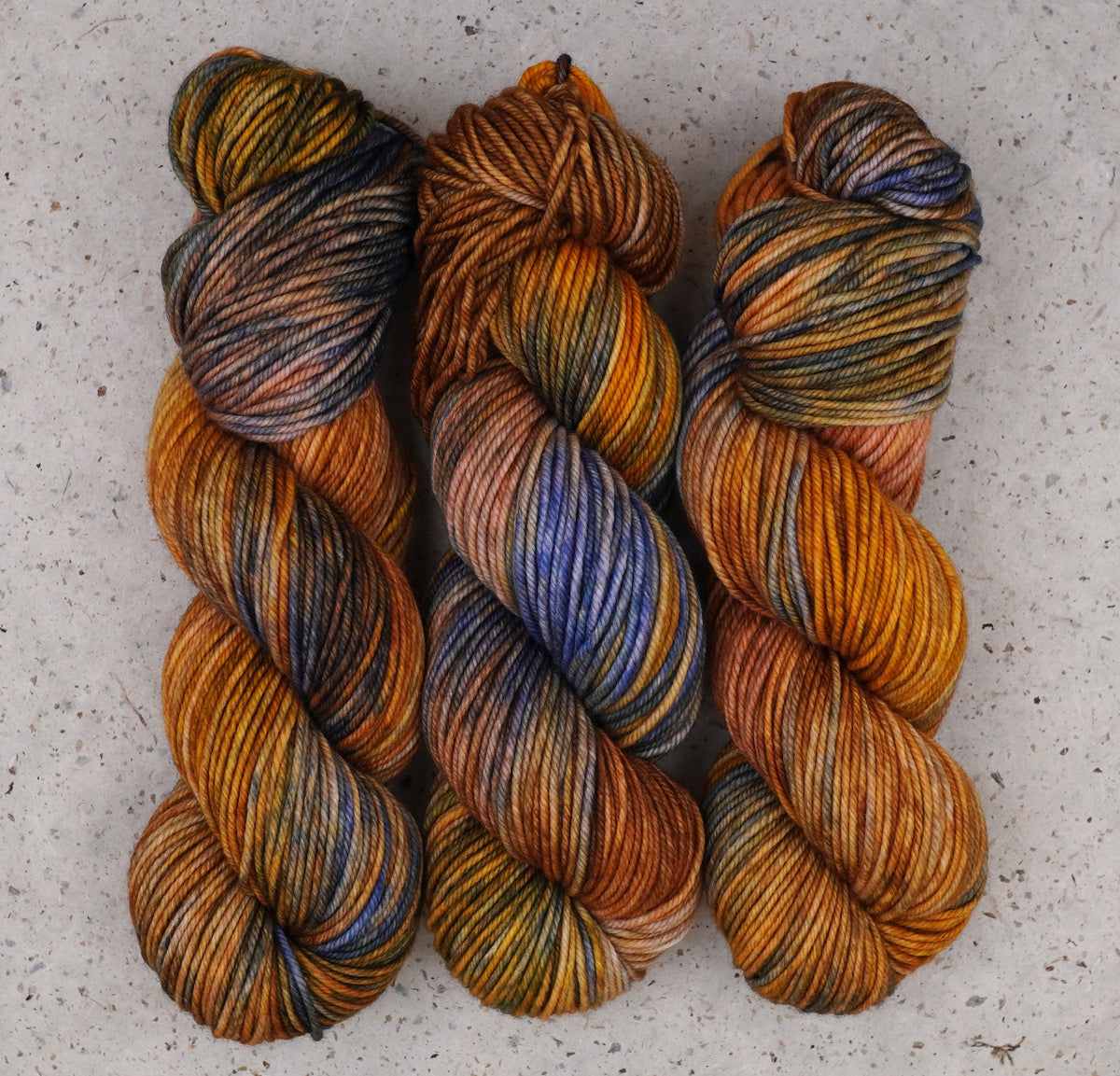 Three twisted hanks of variegated yarn laid flat on a light gray textured surface. The yarn displays a rich color-shifting pattern with warm orange, brown, and gold tones, accented by dark navy and deep blue sections.