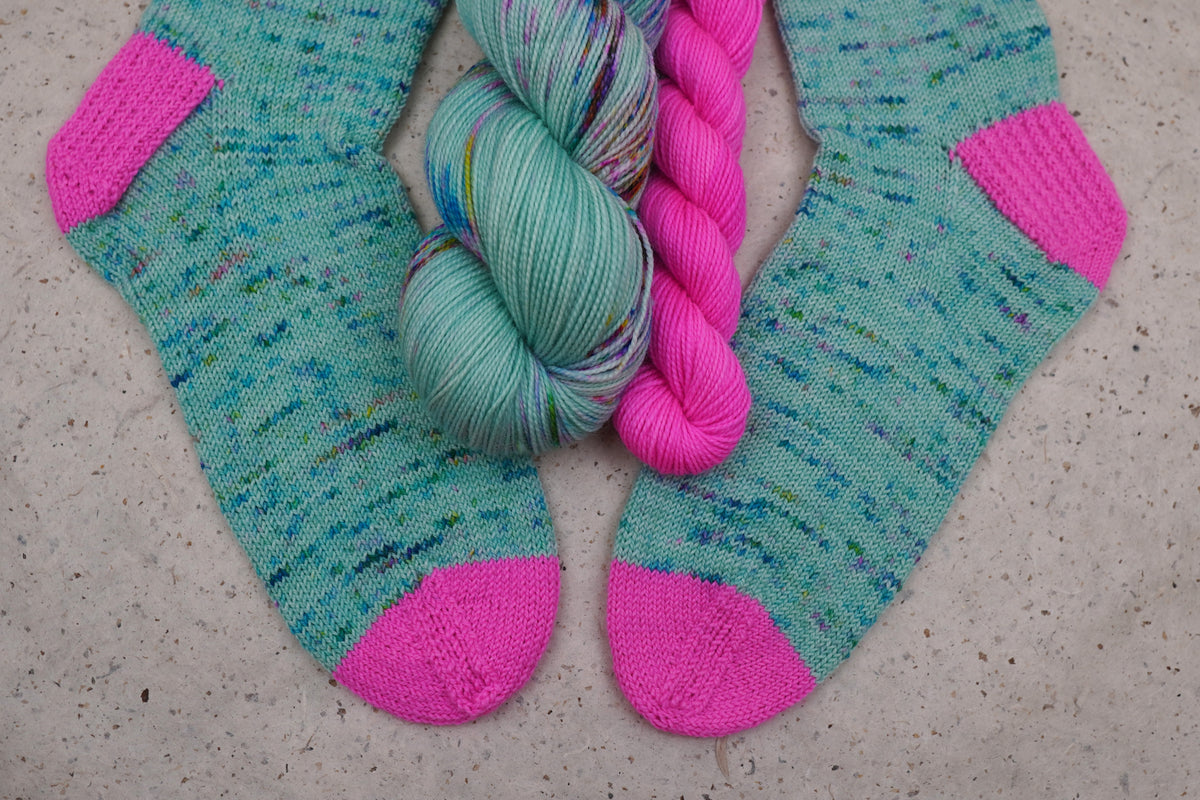 Hand-knit socks in Wild Woollys Undersea Garden with contrasting hot pink heels and toes, shown with the sock set skeins