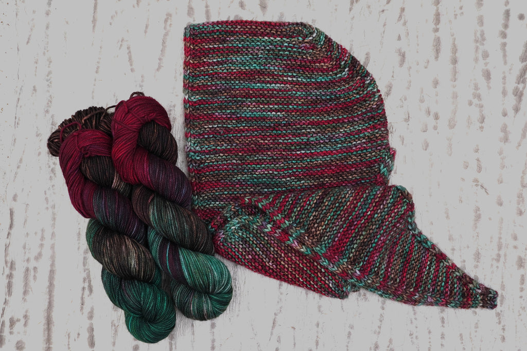 A hand-knit shawl in Wild Woollys Woodland Christmas, shown with three skeins in deep forest green, crimson, and near-black