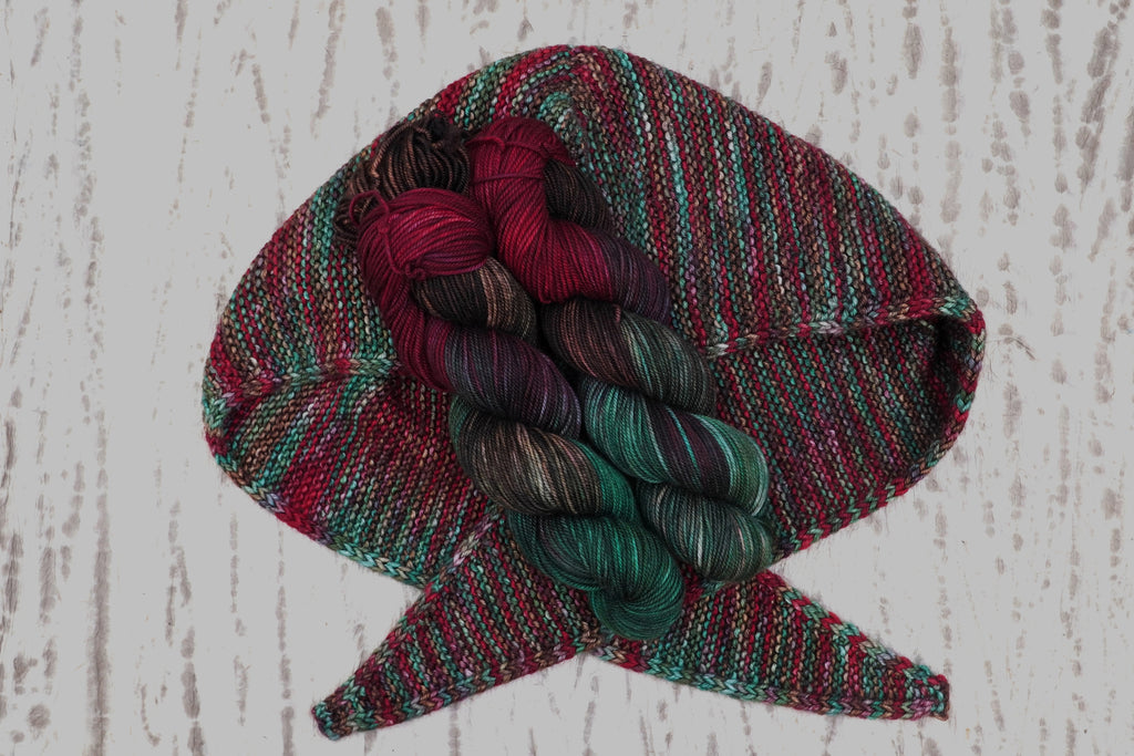 A hand-knit shawl in Wild Woollys Woodland Christmas, shown with three skeins in deep forest green, crimson, and near-black